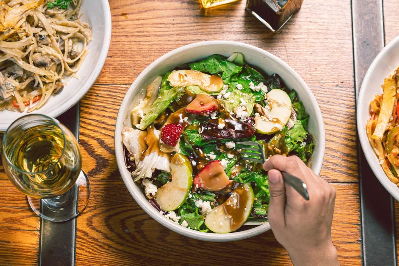 A bowl of pasta salad with sliced apple, greens and pomegranate seeds on a wooden table