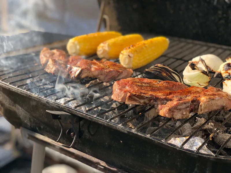Steaks and husked corn cobs cooking side by side over a charcoal grill