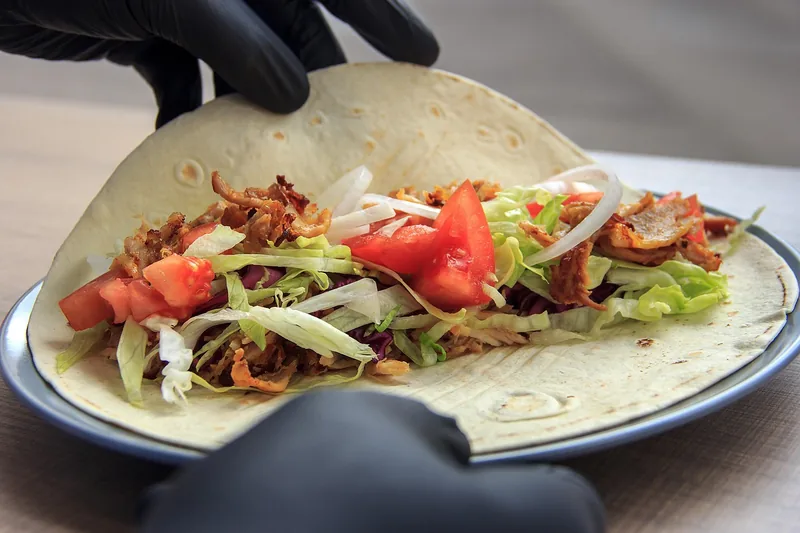 Gloved hands holding a flatbread loaded with seasoned ground beef, shredded lettuce and diced tomato