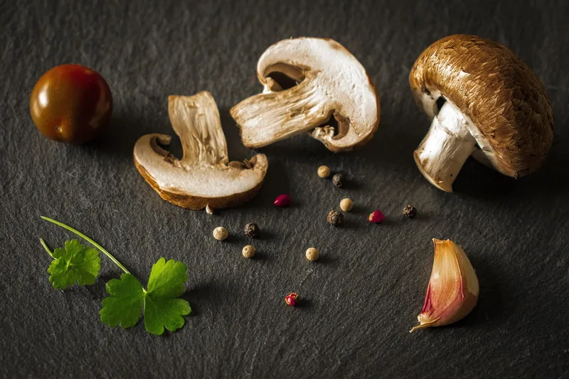 Sliced and whole brown mushrooms on dark slate with garlic and peppercorns