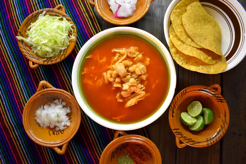 A bowl of chicken tortilla soup with tortilla chips, lime, onion and cabbage around it
