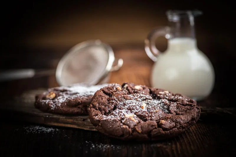 A dark chocolate cookie dusted with powdered sugar on a wooden board, a jug of milk behind it