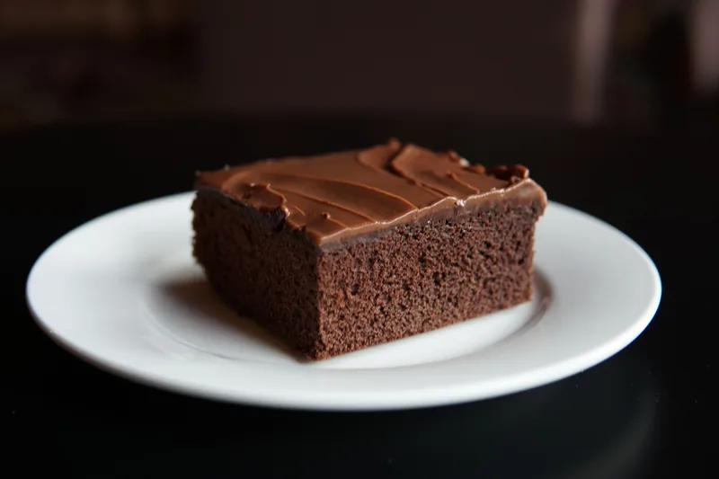 A square slice of frosted chocolate cake on a white plate