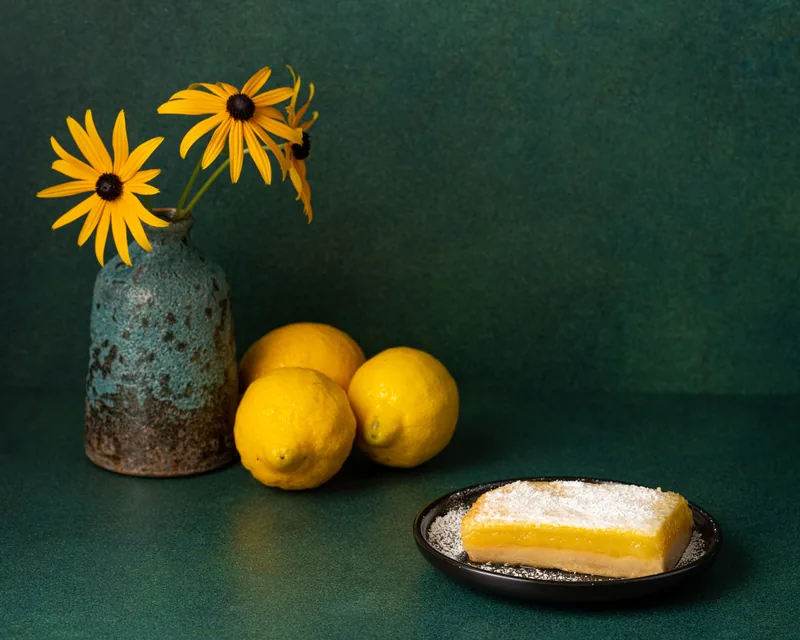 A single lemon bar on a dark plate with lemons and flowers