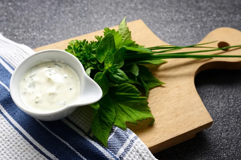 A jug of creamy sauce beside fresh herbs