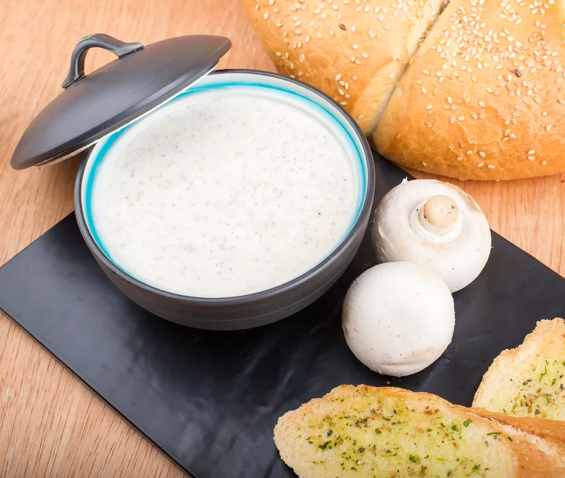 A dark pot of thick creamy mushroom dip with a whole mushroom and garlic bread beside it