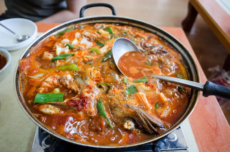 A pan of tomato-based fish stew with pieces of white fish and fresh herbs, a spoon resting in it