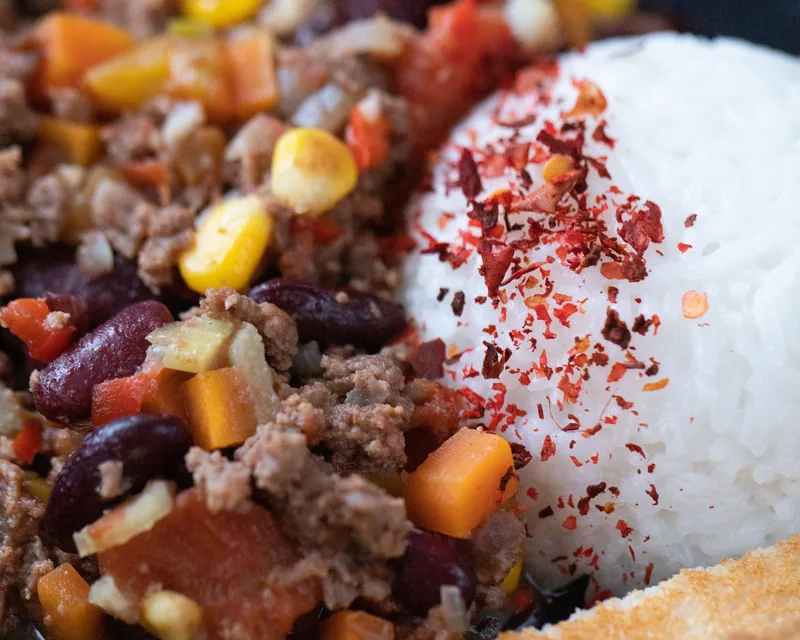 Close-up of chili with ground meat, kidney beans, corn and tomato, rice alongside, dusted with chili flakes