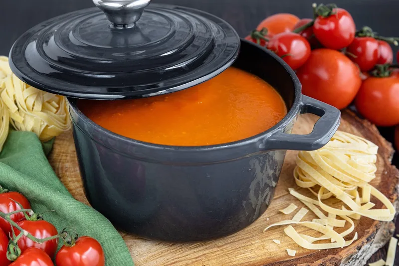 A pot of tomato soup beside dried pasta and fresh tomatoes