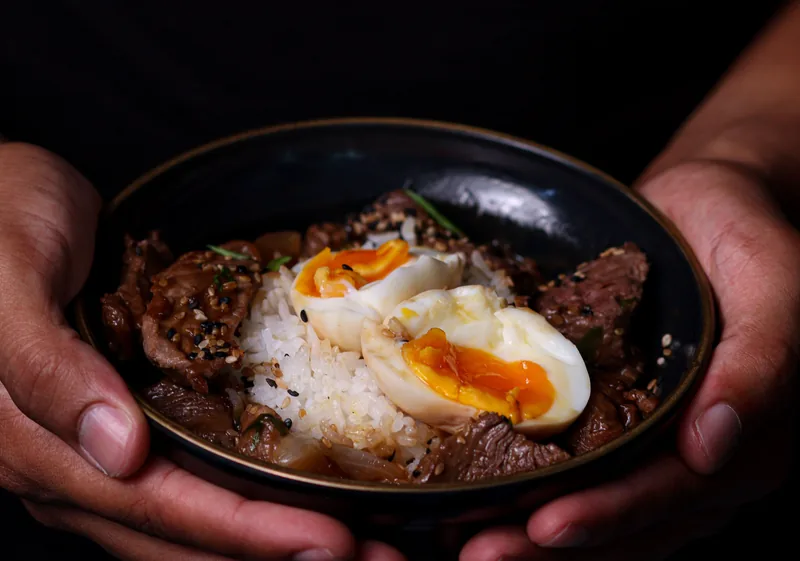 A bowl of rice, beef and a soft egg held in two hands